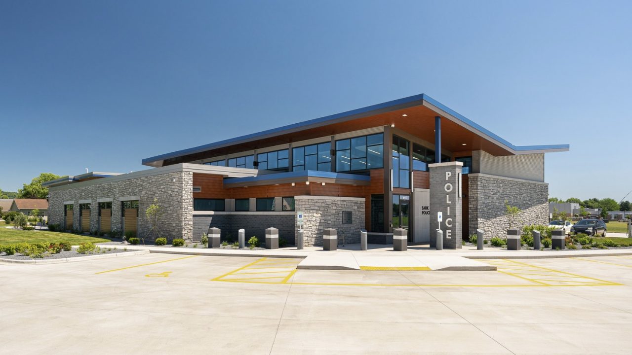 View of contemporary police station facility in Sauk City, Wisconsin with grey stone and wood exterior, ample windows and V-shaped roof with solar array.